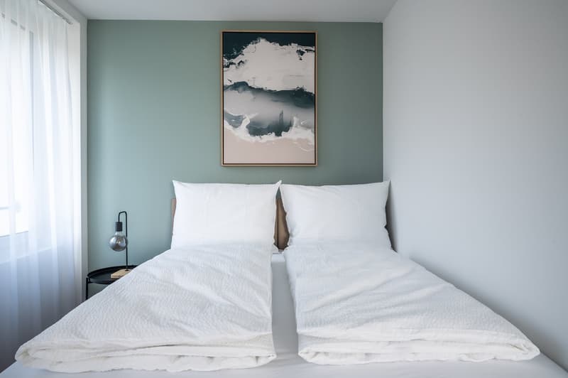 A bedroom with a double bed, white bedding, a nightstand with a lamp, and a framed picture on the wall.
