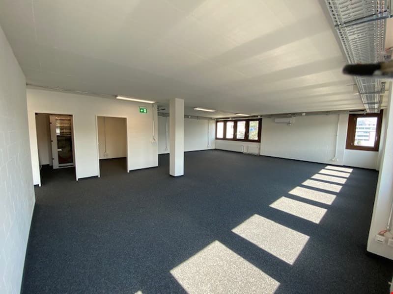 Large open office space with gray carpeted flooring, white walls, and several windows providing natural light. The space appears to be empty and ready for use.