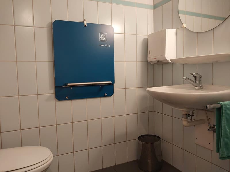 white tiles, sink, faucet, mirror, paper towel dispenser, toilet, waste bin, towel rack