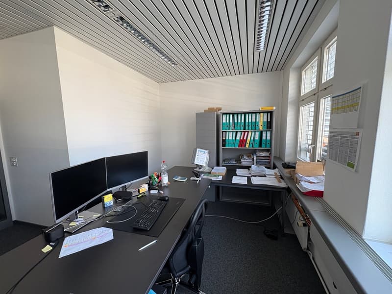 An empty office with two desks, monitors, keyboards, mouse, bottles, papers, and a cabinet with files.