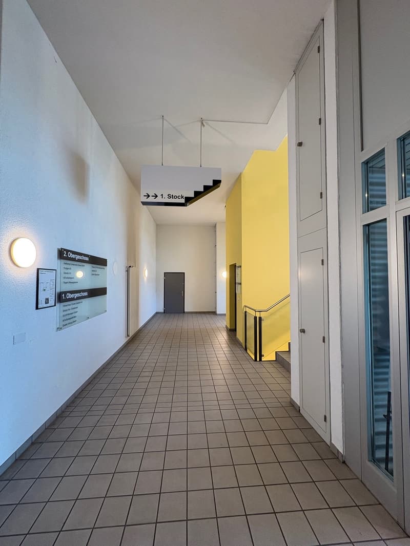 tiled floor, white walls, yellow accent wall, two floors, signs indicating floor levels, ceiling lights, doors, and a staircase