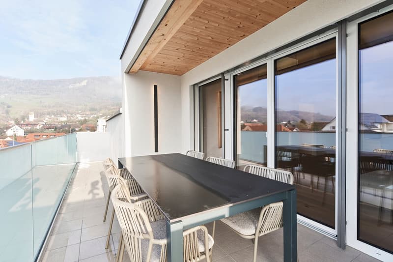 Spacious balcony with a large glass sliding door, wooden ceiling, and a dining table with chairs overlooking a scenic view of the surrounding town and mountains