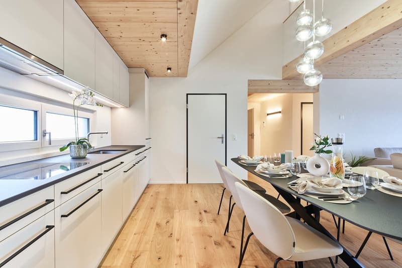 The image shows a modern, open-concept kitchen and dining area. The kitchen features white cabinets, a black countertop, and a dishwasher. The dining area has a large wooden table with white chairs. The ceiling has exposed wooden beams and recessed lighti