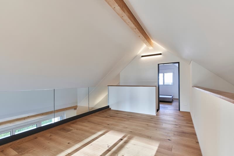 Spacious attic bedroom with hardwood floors, white walls, and exposed wooden beams. The room features a glass railing overlooking the lower level, providing a balcony-like feel. There is a window allowing natural light to enter the space.