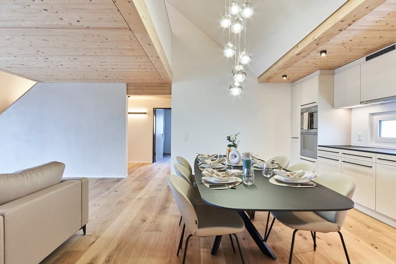 The image shows an open concept living area with a dining table and chairs, a kitchen visible in the background, and a wooden ceiling with recessed lighting. The floors are hardwood, and there is a sofa visible in the living room area. The kitchen appears