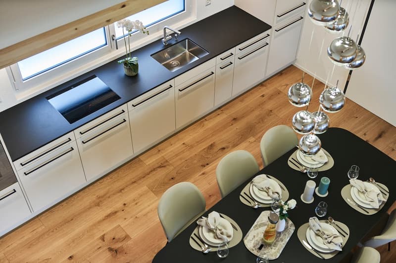Modern kitchen with white cabinets, black countertop, stainless steel sink, and hardwood floor. Dining table with place settings and decorative elements.