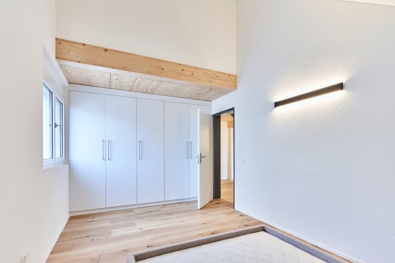 Spacious bedroom with white built-in wardrobes, wooden ceiling beams, and hardwood flooring. The room has a bright and airy feel with a large window, suggesting the possibility of a balcony or terrace.