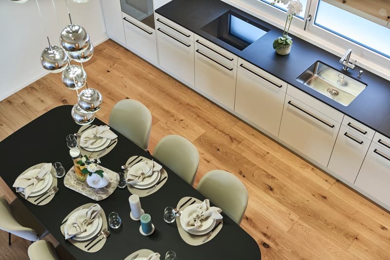 Modern kitchen with dark countertops, light cabinets, and hardwood floors. Dining table with place settings and decorative elements. Dishwasher visible in the kitchen cabinetry.