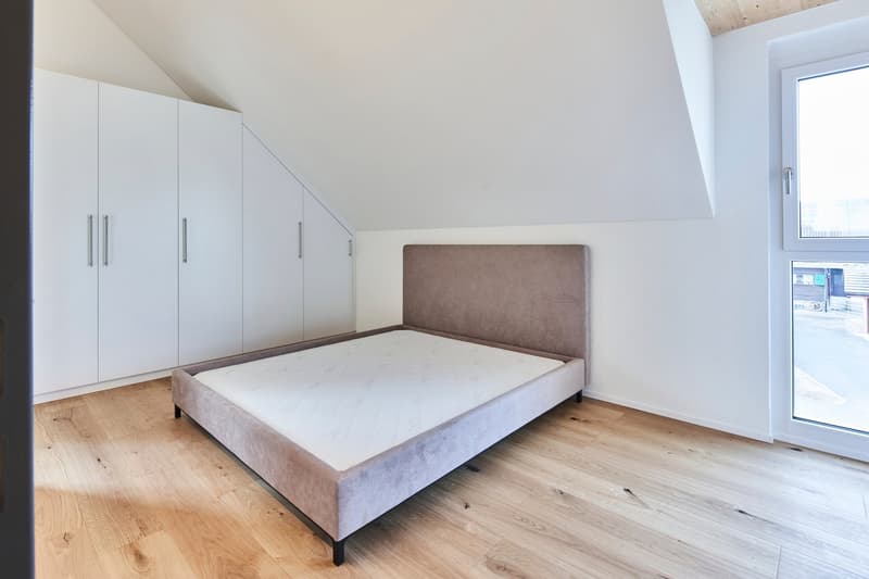 Spacious bedroom with white built-in wardrobes, a gray upholstered bed frame, and hardwood flooring. Large window provides natural light.