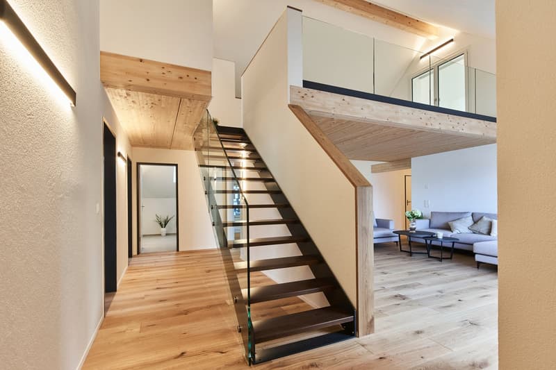 The image shows a modern and open-concept interior space with a wooden staircase leading to an upper level. The staircase features glass panels and wooden steps, creating a sleek and minimalist design. The walls are painted in a light color, and the floor