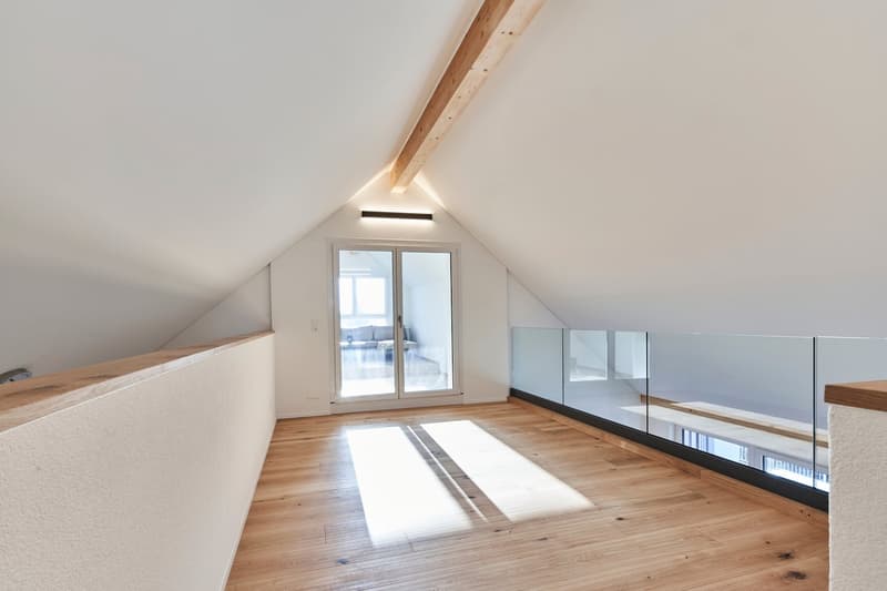 Spacious attic or loft space with wooden floors, slanted ceiling, large windows, and glass railing overlooking the lower level.