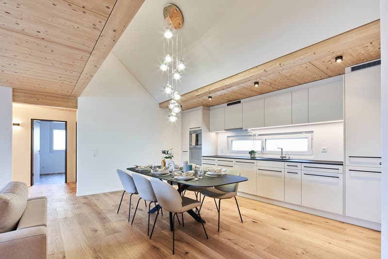 Open-concept kitchen and dining area with high ceilings, wooden beams, and modern lighting fixtures. The kitchen features white cabinets, stainless steel appliances, and a large dining table with chairs. The flooring is hardwood throughout.
