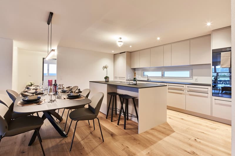 Open-concept kitchen and dining area with modern, light-colored cabinets, a kitchen island with bar stools, a dining table with chairs, and hardwood flooring. The space features recessed lighting and large windows providing natural light.