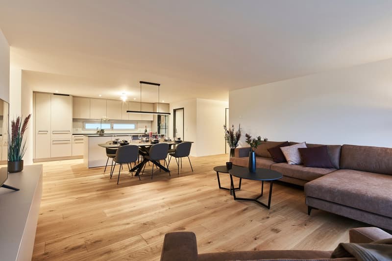 The image shows an open concept living room and kitchen area with hardwood floors, a large L-shaped sofa, a dining table with chairs, and a modern kitchen with white cabinets and a dishwasher visible.