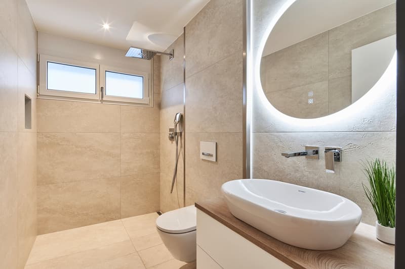 Modern bathroom with beige tile walls, a large oval-shaped sink, a toilet, and a walk-in shower with glass enclosure. The bathroom has large windows providing natural light, and features a round backlit mirror above the sink.