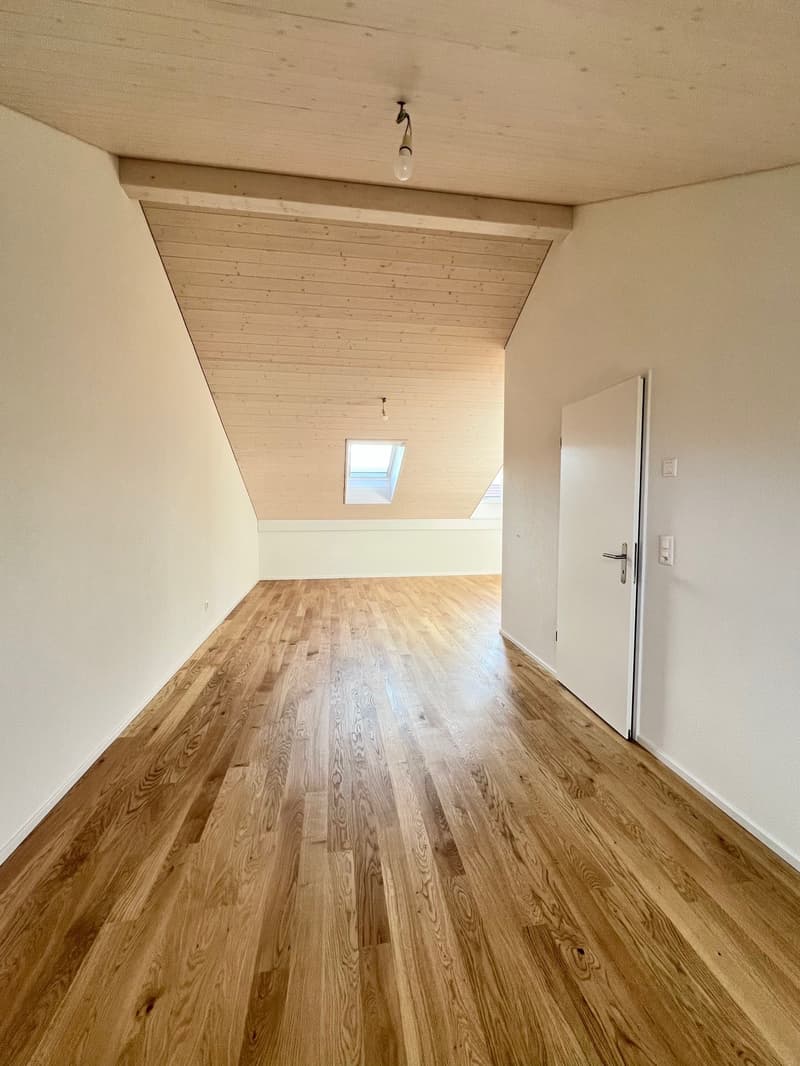 Empty room with wooden floors, white walls, a wooden ceiling, an open attic window, and a white door with a handle.