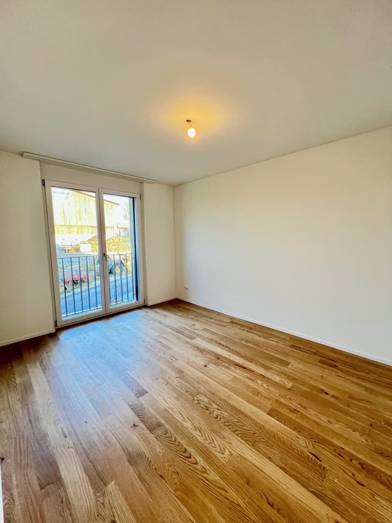 Empty room, wooden floors, sliding glass doors leading to balcony, white walls, large window