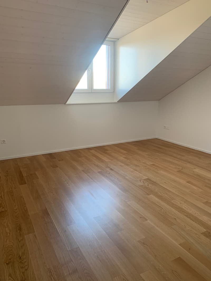 Spacious bedroom with hardwood flooring, white walls, and a large window providing natural light.