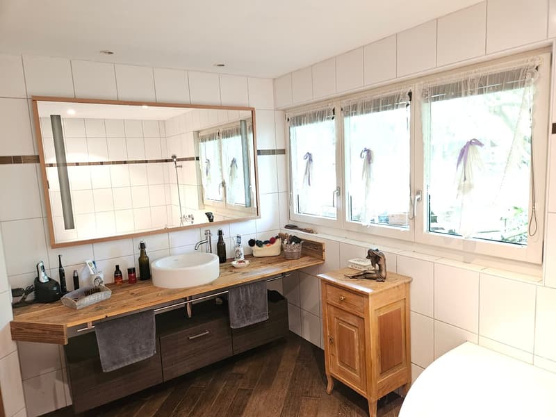 Bathroom with wooden floor, white tiles, large mirror, wooden vanity, white sink, wooden cabinet, white bathtub, wooden drawer, towels, and various toiletries.