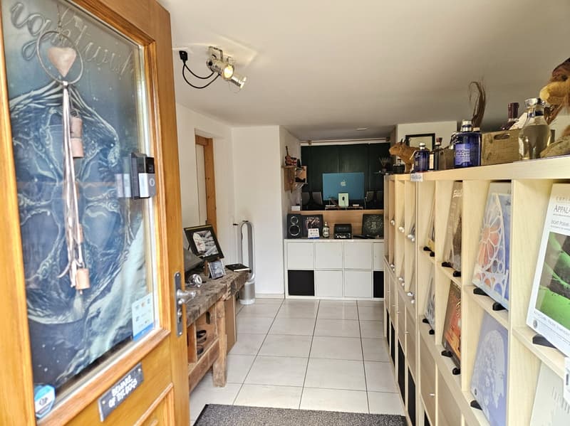 A room with a wooden door, a wooden desk, a white tiled floor, a wooden cabinet, a shelf with books and items, a monitor, a speaker, and a lamp.