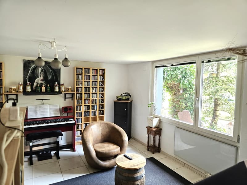 A room with a piano, a bookshelf, a chair, a table, a radiator, and a window with a view of trees outside