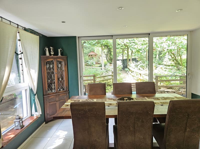 Dining room with large windows, wooden table, brown chairs, wooden cabinet, white curtains, tiled floor, view of garden