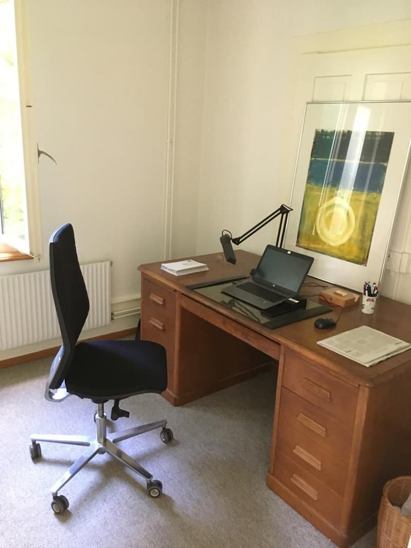 The image shows a home office setup with a wooden desk, a laptop computer, and a black office chair. There is also a framed artwork on the wall and some other office supplies on the desk.