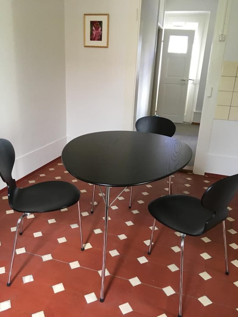 The image shows a dining area with a round black table and four black chairs. The floor is tiled with a red and white pattern. There is a framed artwork on the wall and a doorway leading to another room visible in the background.