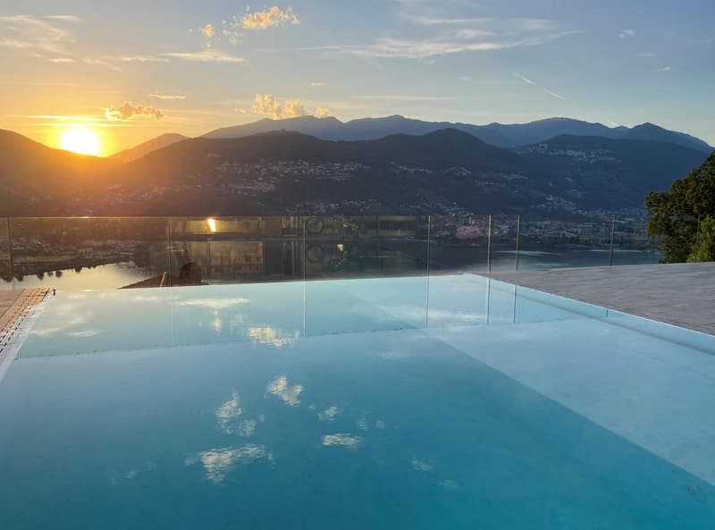 Stunning infinity pool overlooking a mountainous landscape at sunset. The pool has a glass barrier, providing an unobstructed view of the surrounding hills and valleys. The sky is filled with warm, golden hues as the sun dips below the horizon.