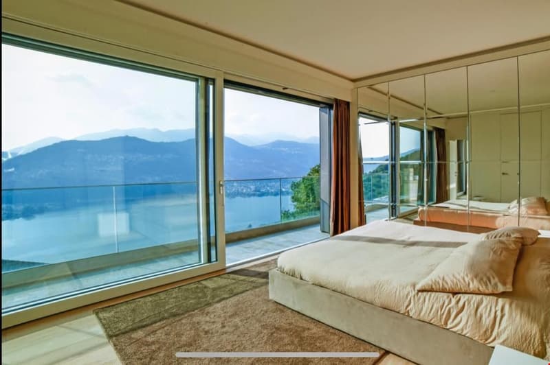 Spacious bedroom with large windows offering a panoramic view of the mountains and lake. The room features a king-size bed, plush carpeting, and a minimalist design aesthetic.