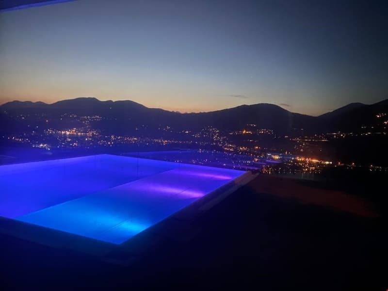 Nighttime view of a swimming pool overlooking a mountainous landscape with a city in the distance. The pool is illuminated with blue and purple lighting, creating a serene and picturesque scene.