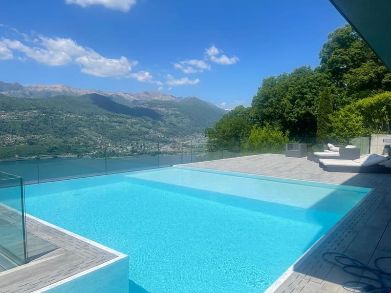 The image shows a large outdoor swimming pool with a stunning view of the surrounding mountains and lush greenery. The pool has a glass railing and is surrounded by a wooden deck with lounge chairs. The sky is a beautiful blue with fluffy white clouds, cr