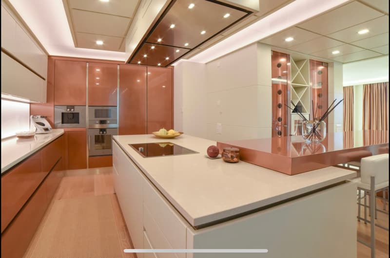 Spacious modern kitchen with sleek cabinetry in a warm orange-brown tone, stainless steel appliances including a dishwasher, and a large central island with a white countertop. The kitchen has recessed lighting and an open layout that connects to the adja