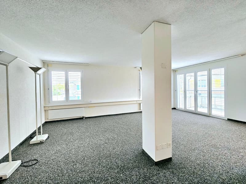 An empty room with carpeted floor, white walls, ceiling lights, glass sliding doors, windows with shutters, central column with electrical outlets, and floor lamps.