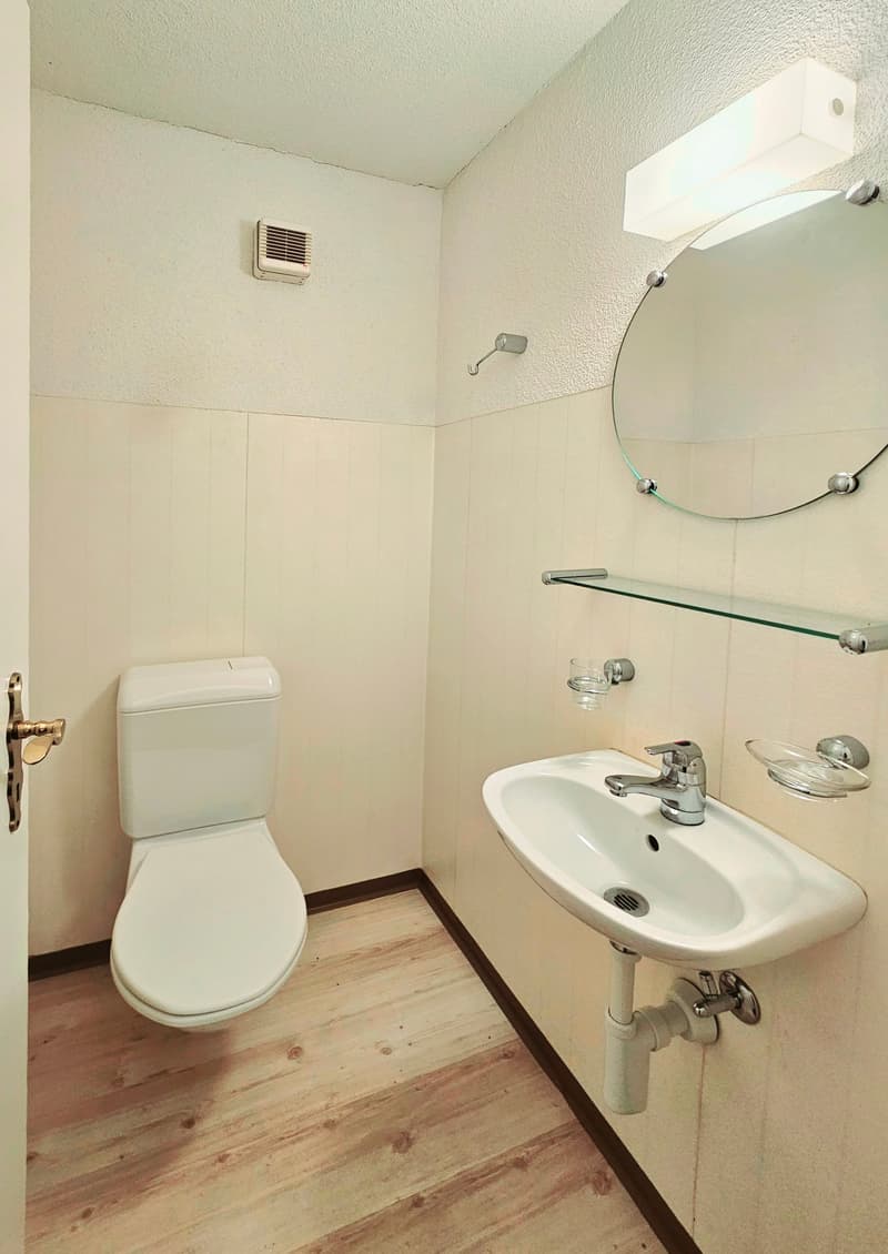 A small, white-tiled bathroom with a toilet, sink, and round mirror above the sink.