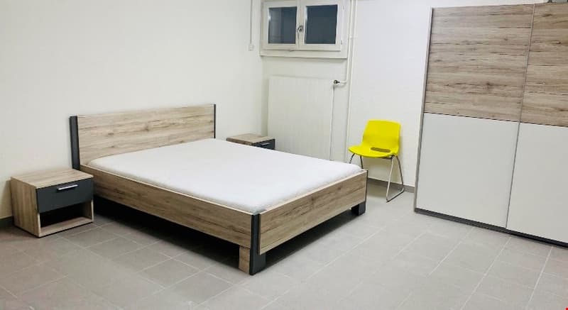 The room has a bed, two nightstands, a chair, and a wardrobe. The bed is made with white sheets, and the nightstands have drawers. The chair is yellow, and the wardrobe is white and brown. The walls are white, and there is a window with a white frame.