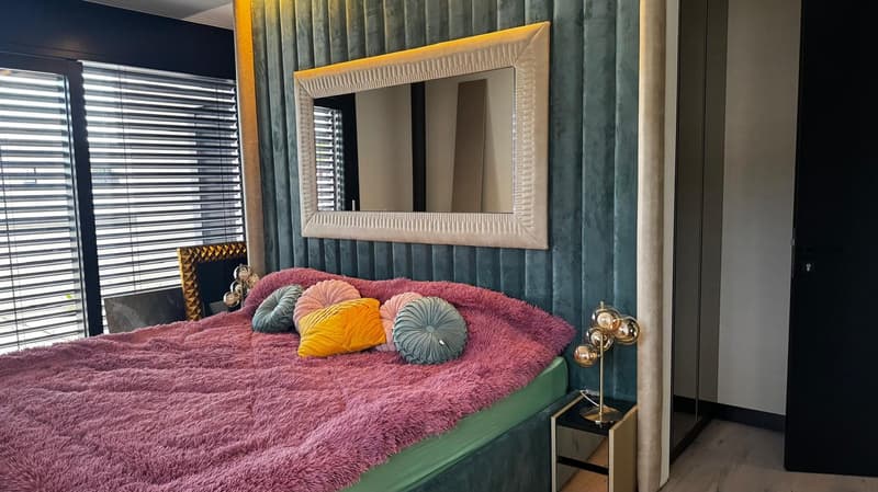 Large bed with pink blanket and pillows, a table with lamp, a mirror on the wall and large windows with blinds