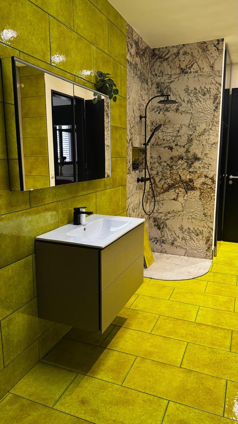 Bathroom with yellow tiled walls, green wall tiles, modern design vanity, white porcelain sink, mirror, shower area with marble-like wall, shower head, and yellow tile flooring.