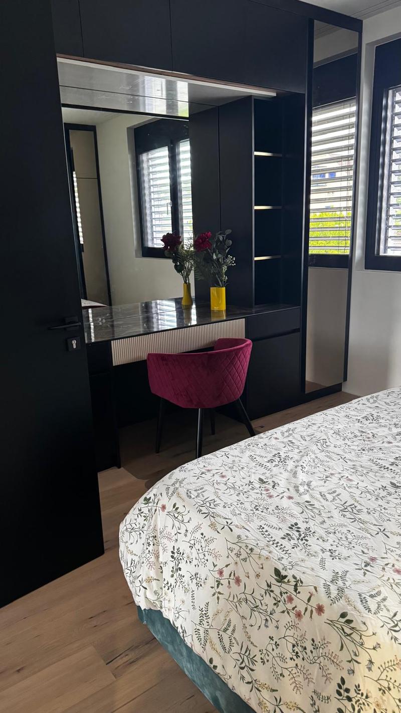 wooden floor, a large bed, desk with mirror, purple chair, window with blinds, flower vase