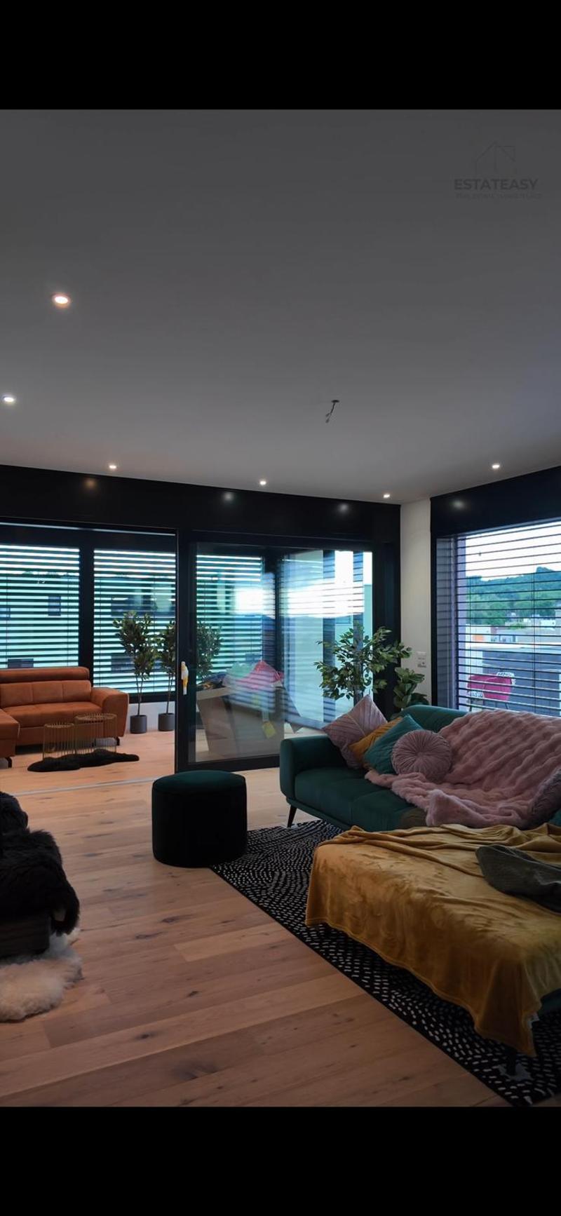 Living room with open floor plan, large windows, modern decor, wooden flooring, glass doors, potted plants, white and green sofa with throw pillows, black rug, ottoman, decorative items, indoor plants