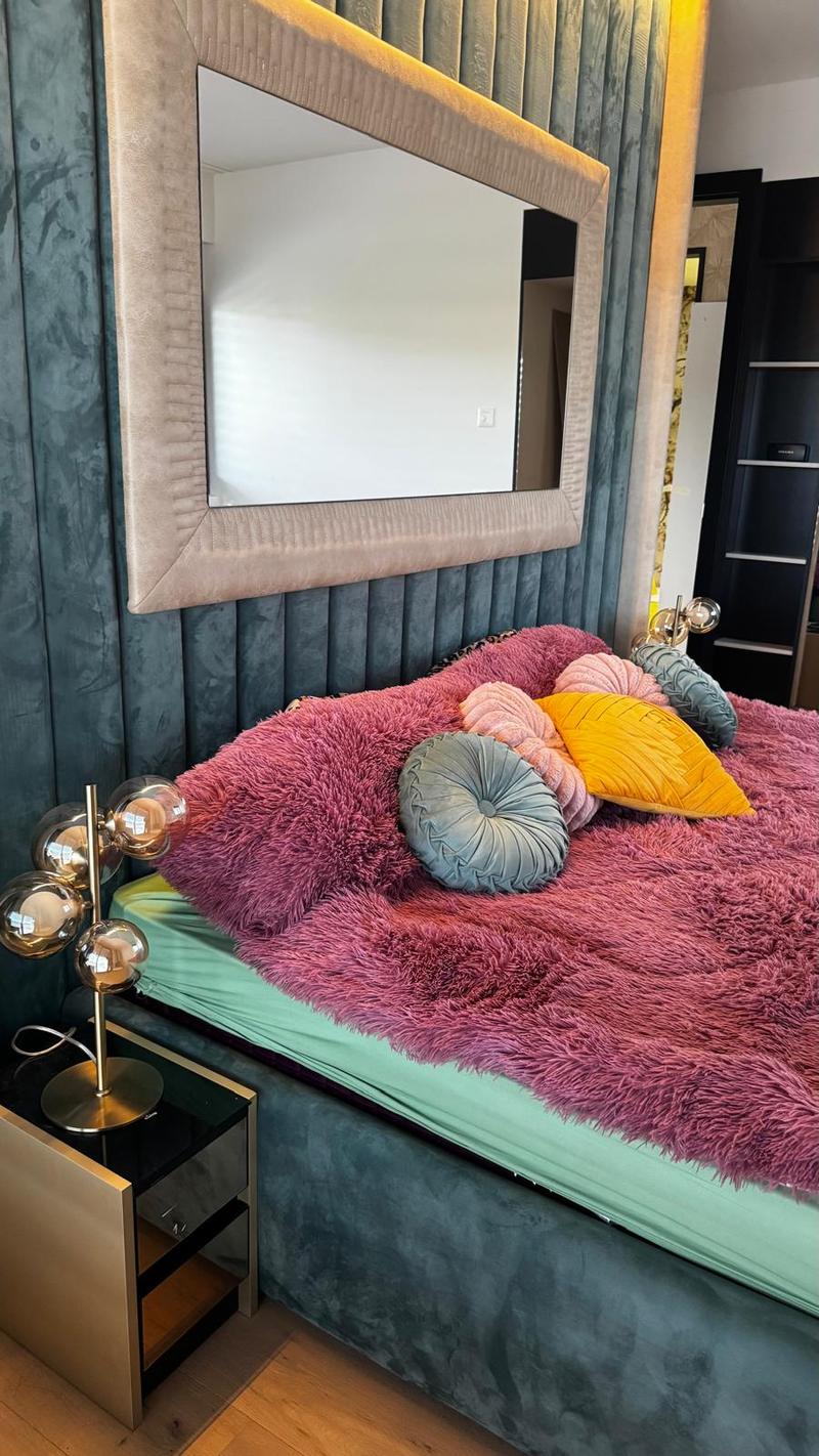 Bedroom with a bed with pink blankets, pink and yellow pillows, a lamp, mirror, dark blue walls.