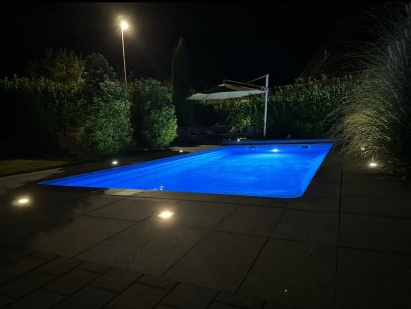 Swimming pool at night, illuminated lights, small umbrella, shrubbery, pathway, bushes, and greenery