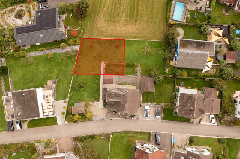 Plot of land marked with a red square, houses on sides, greenery, driveways, and cars parked in front.
