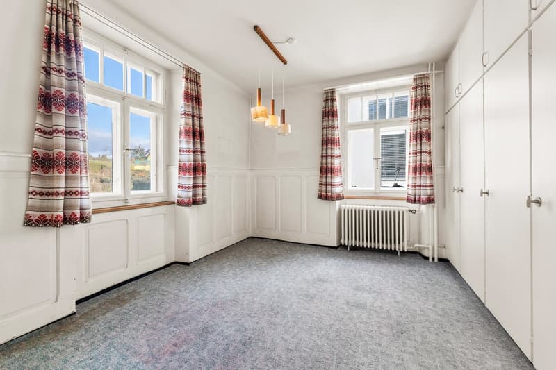 Large empty room, white walls, wooden ceiling, 3 windows with curtains, radiator, carpeted floor