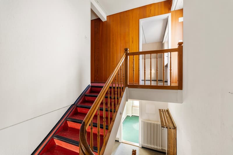House with wooden staircase and wooden railing, red steps, white walls, wooden floor, and white radiator.