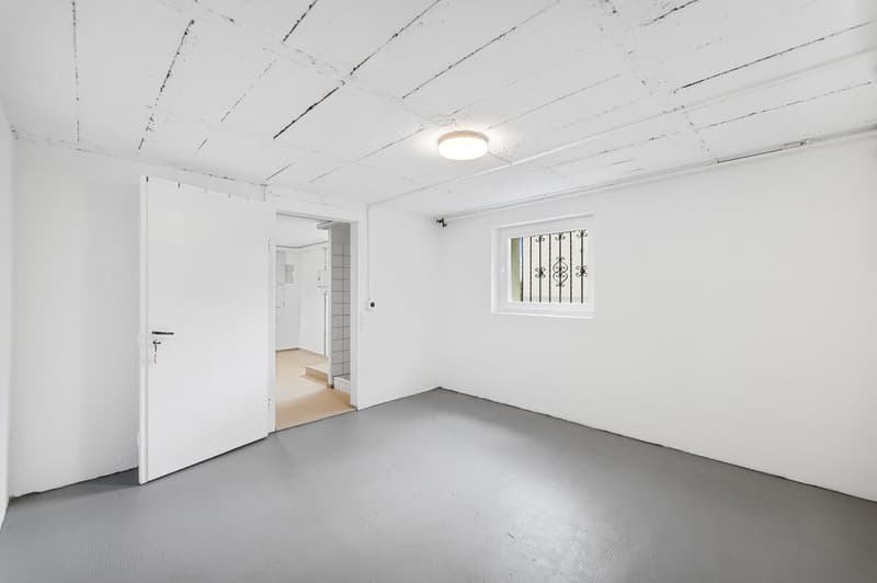 Empty room with white walls, gray floor, open door, small window with metal grill, ceiling light