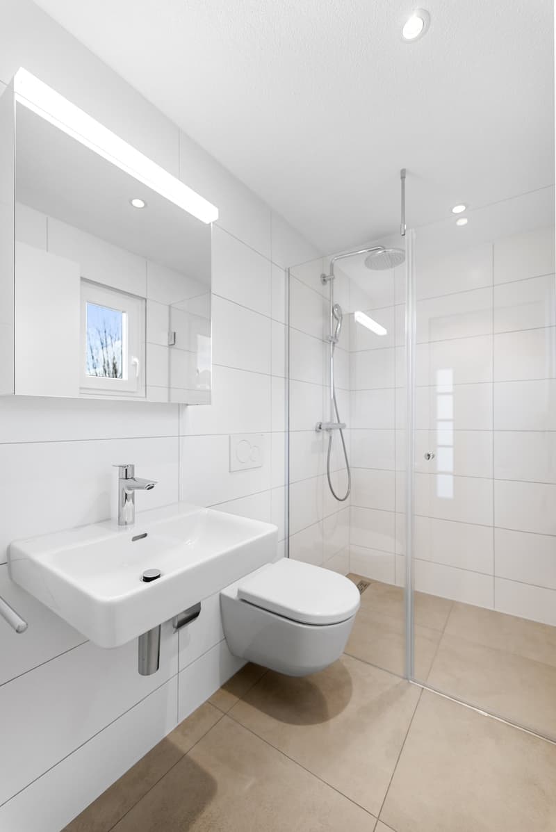 modern bathroom with white tiles, glass shower enclosure, white sink, silver faucet, white toilet, mirror with light