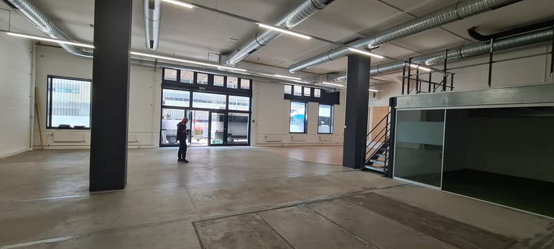 Large open commercial space with concrete floors, exposed ductwork, and large windows. The space appears to be an unfinished or partially renovated commercial property, with a person visible in the background.