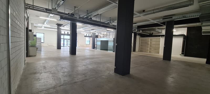 Large open commercial space with concrete floors, exposed ceiling ducts, and black support columns. The space appears to be unfinished or under renovation, with a few appliances or equipment visible. The windows provide natural light to the space.