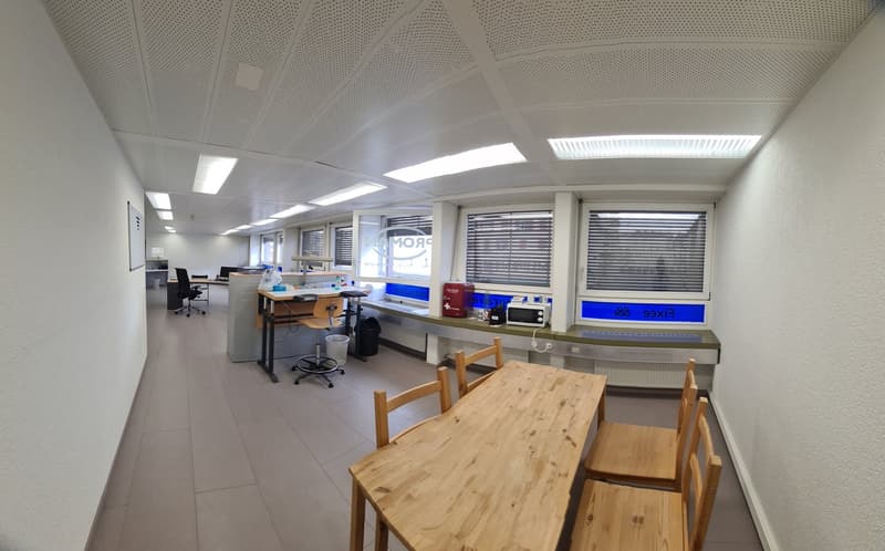 Open office space with cubicles, desks, and chairs. Wooden table and chairs in the foreground. Shelves and equipment visible along the walls.